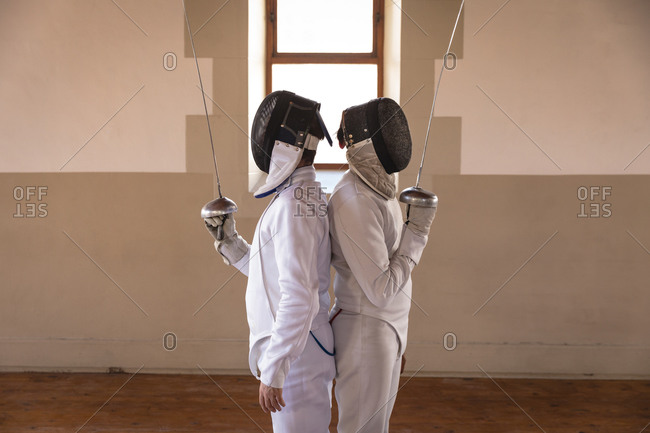 Caucasian and a mixed race sportsmen wearing protective fencing outfit during a fencing training session, standing back to back, holding epees in front of faces. Fencers training at a gym.