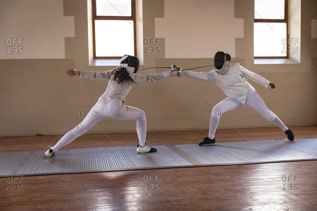 Caucasian and African American sportswomen wearing protective fencing outfits during a fencing training session, taking aim at each other with their epees. Fencers training at gym.
