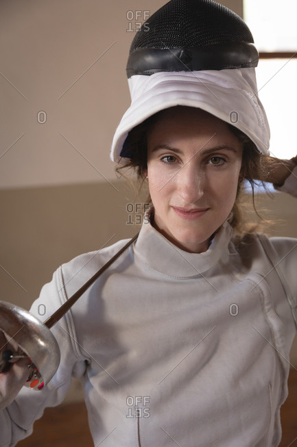 Portrait of Caucasian sportswoman wearing protective fencing outfit during a fencing training session, looking at camera with her mask raised and smiling, holding an epee. Fencers training at a gym.
