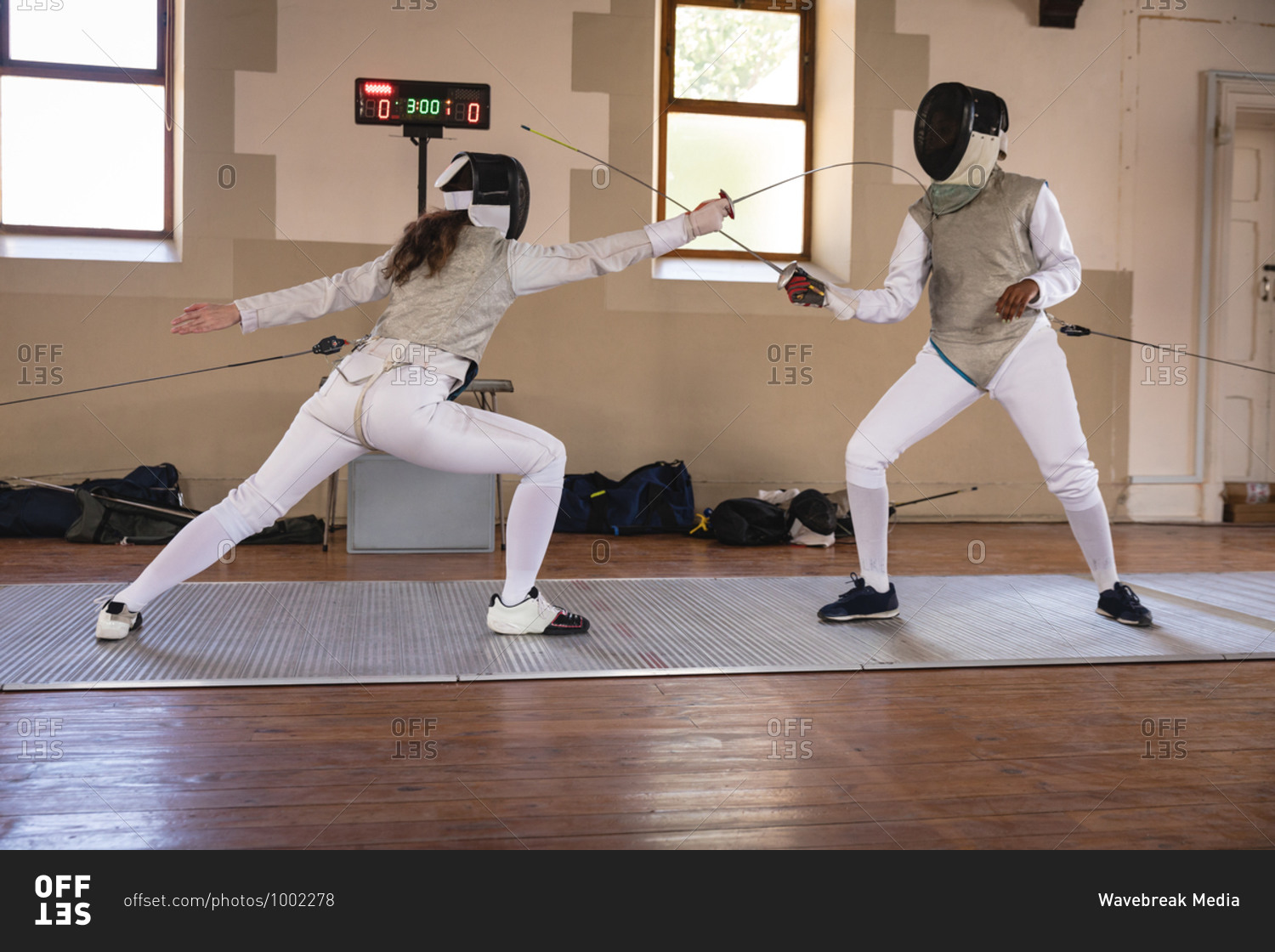 Caucasian and African American sportswomen wearing protective fencing outfits during a fencing training session, jumping taking aim at each other with their epees. Fencers training at gym.
