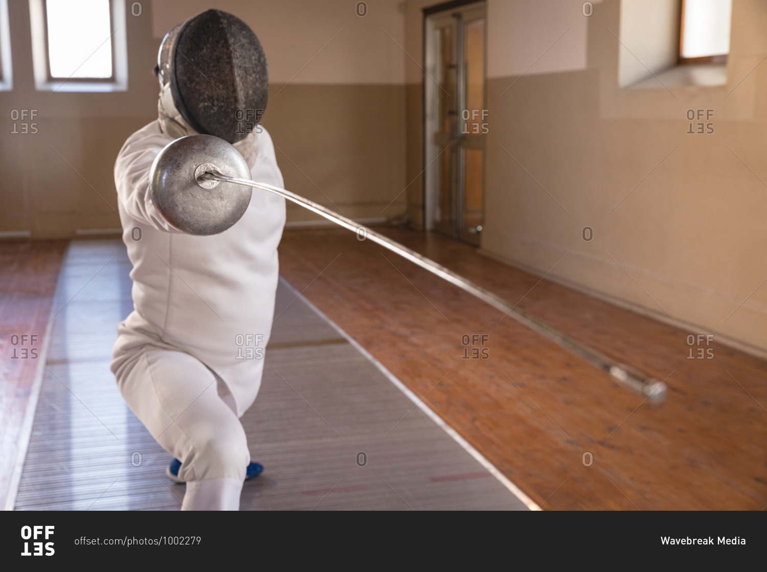 Caucasian sportsman wearing protective fencing outfit during a fencing training session, preparing for a duel, holding an epee and lunging. Fencers training at a gym.