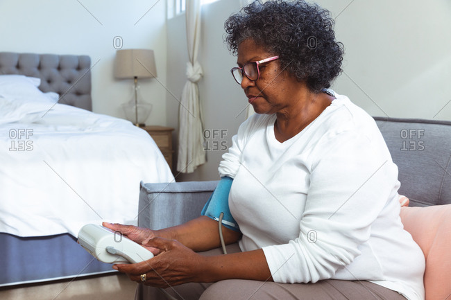 Senior mixed race woman spending time at home, sitting on a chair and taking her blood pressure with a tonometer, social distancing and self isolation in quarantine lockdown