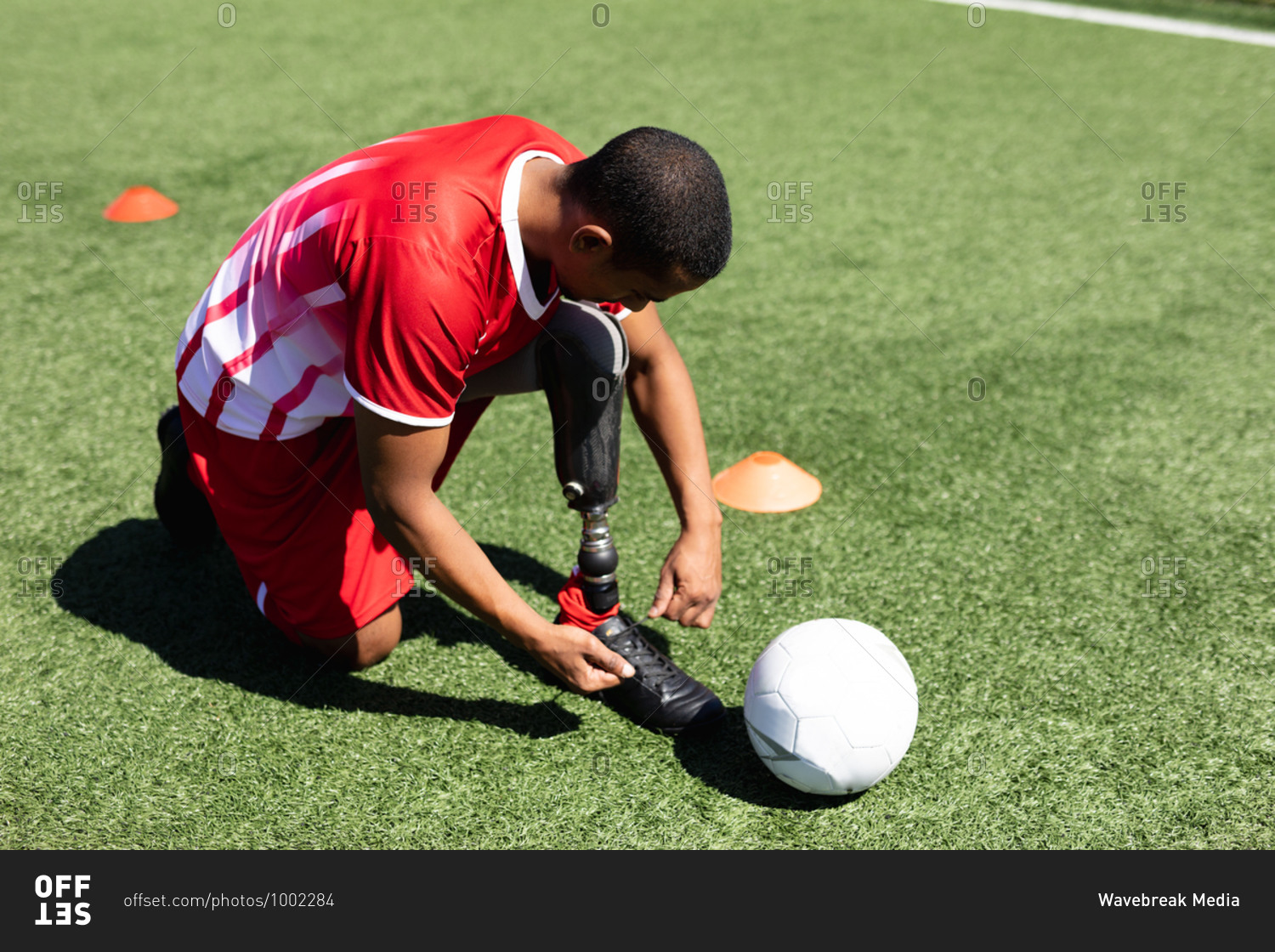 Mixed race male football player with prosthetic leg wearing a team strip training at a sports field in the sun, tying shoelaces ball next to him.