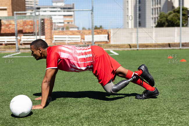 Mixed race male football player with prosthetic leg wearing a team strip training at a sports field in the sun, warming up doing push ups with ball next to him.