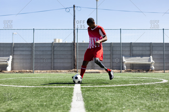 Mixed race male football player with prosthetic leg wearing a team strip training at a sports field in the sun, warming up kicking ball.