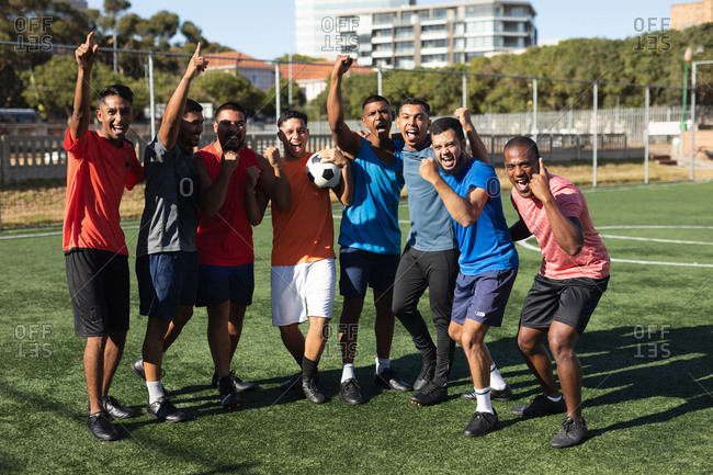 Multi ethnic group of male five a side football players wearing sports clothes training at a sports field in the sun, celebrating victory holding a ball.