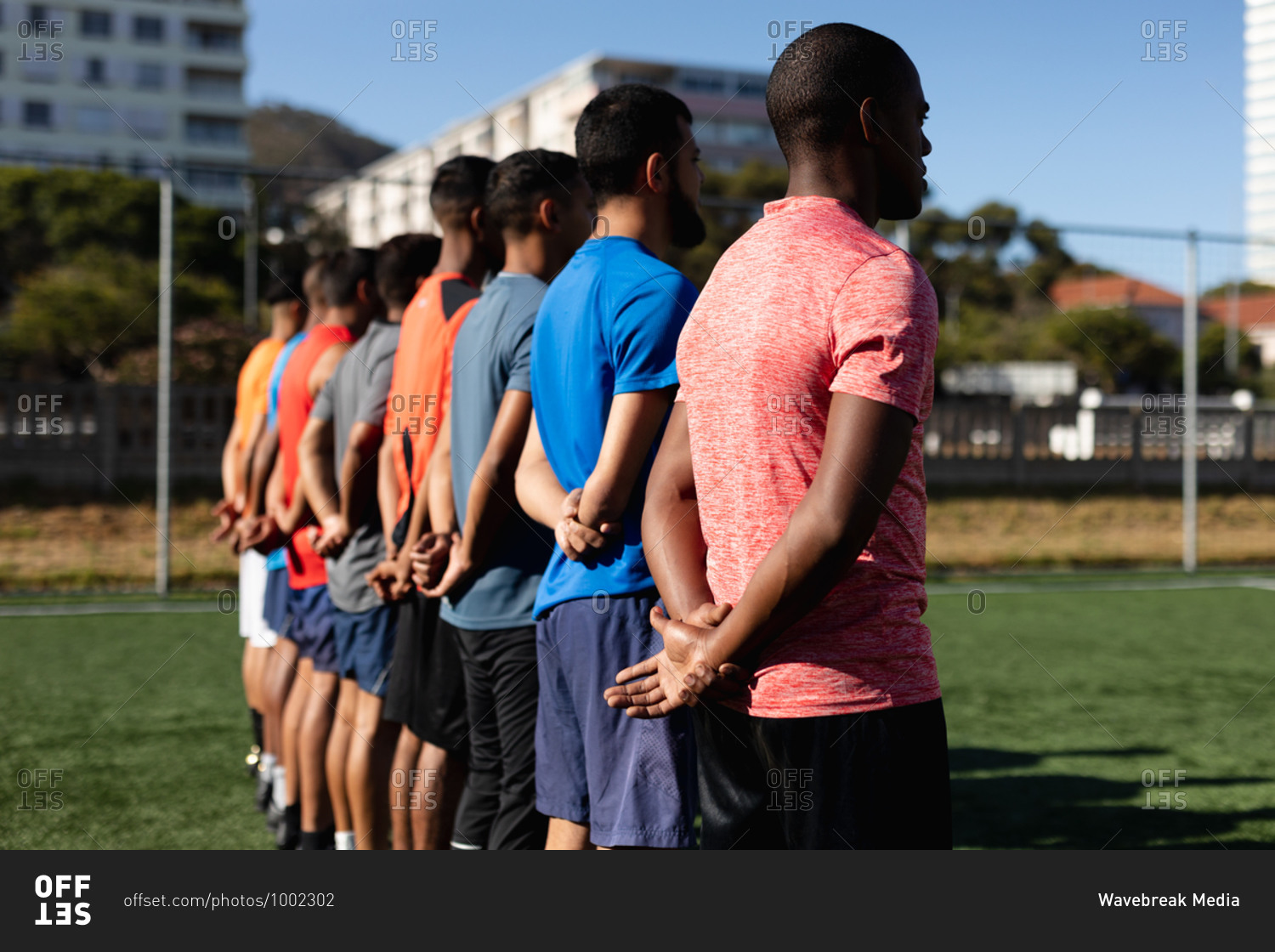 Multi ethnic group of male five a side football players wearing sports clothes training at a sports field in the sun, standing in a row before a game.