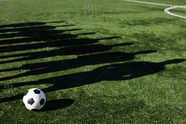 Shadow on grass pitch of group of male football players training at a sports field in the sun, standing next to each other ball next to them.