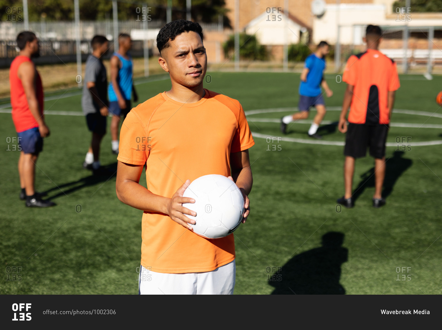Portrait of focused mixed race male five a side football player wearing sports clothes training at a sports field in the sun, holding a ball his teammates warming up in the background.