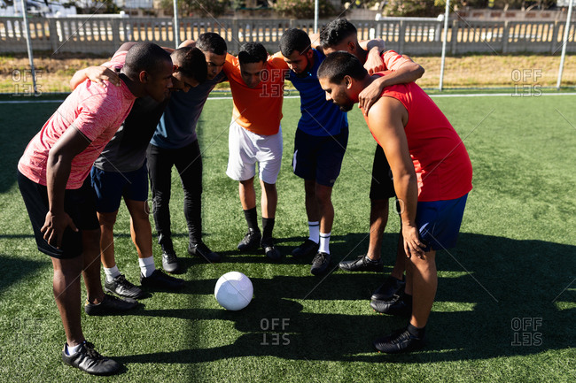 Multi ethnic group of male five a side football players wearing sports clothes training at a sports field in the sun, standing in huddle motivating before a game ball in the middle.