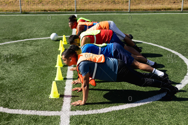 Multi ethnic group of male five a side football players wearing sports clothes and vests training at a sports field in the sun, warming up doing push ups in a row with ball and cones next to them.