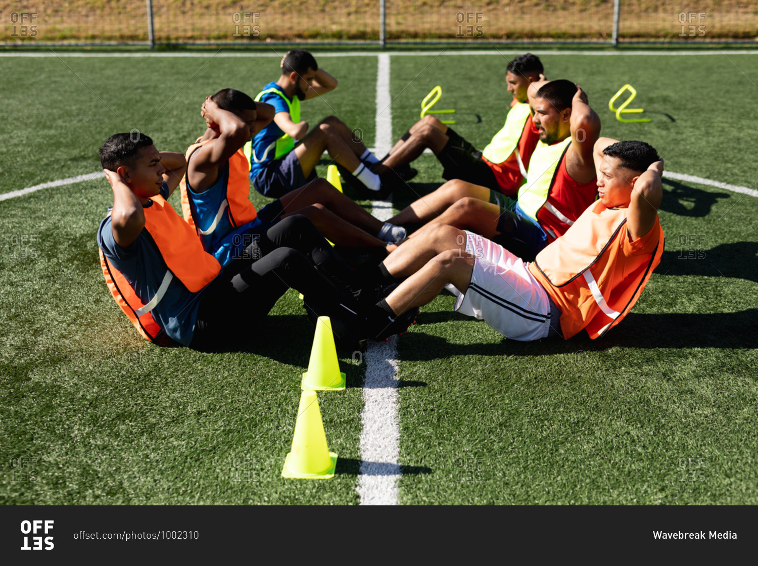Multi ethnic group of male five a side football players wearing sports clothes and vests training at a sports field in the sun, warming up doing sit ups with cones next to them.