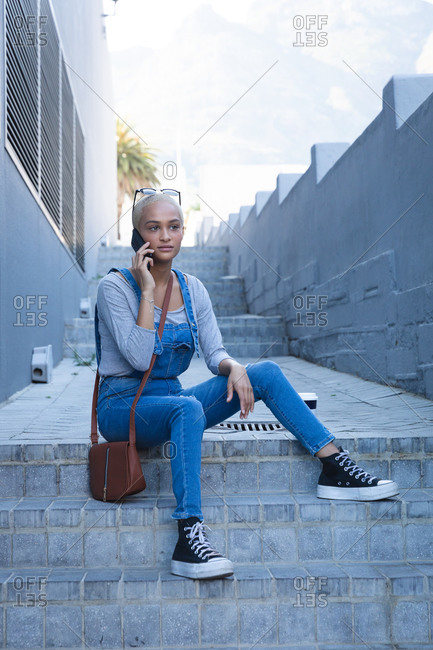 Mixed race alternative woman with short blonde hair out and about in the city on a sunny day, wearing sunglasses and denim dungarees, sitting on steps using smartphone. Urban digital nomad on the go.