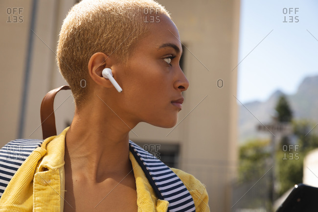 Mixed race alternative woman with short blonde hair out and about in the city on a sunny day, wearing wireless earphones and a backpack, looking away. Urban digital nomad on the go.