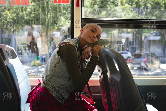 Mixed race alternative woman with short blonde hair out and about in the city, sitting on a bus, leaning on the seat in front, looking away and smiling. Independent urban nomad on the go.
