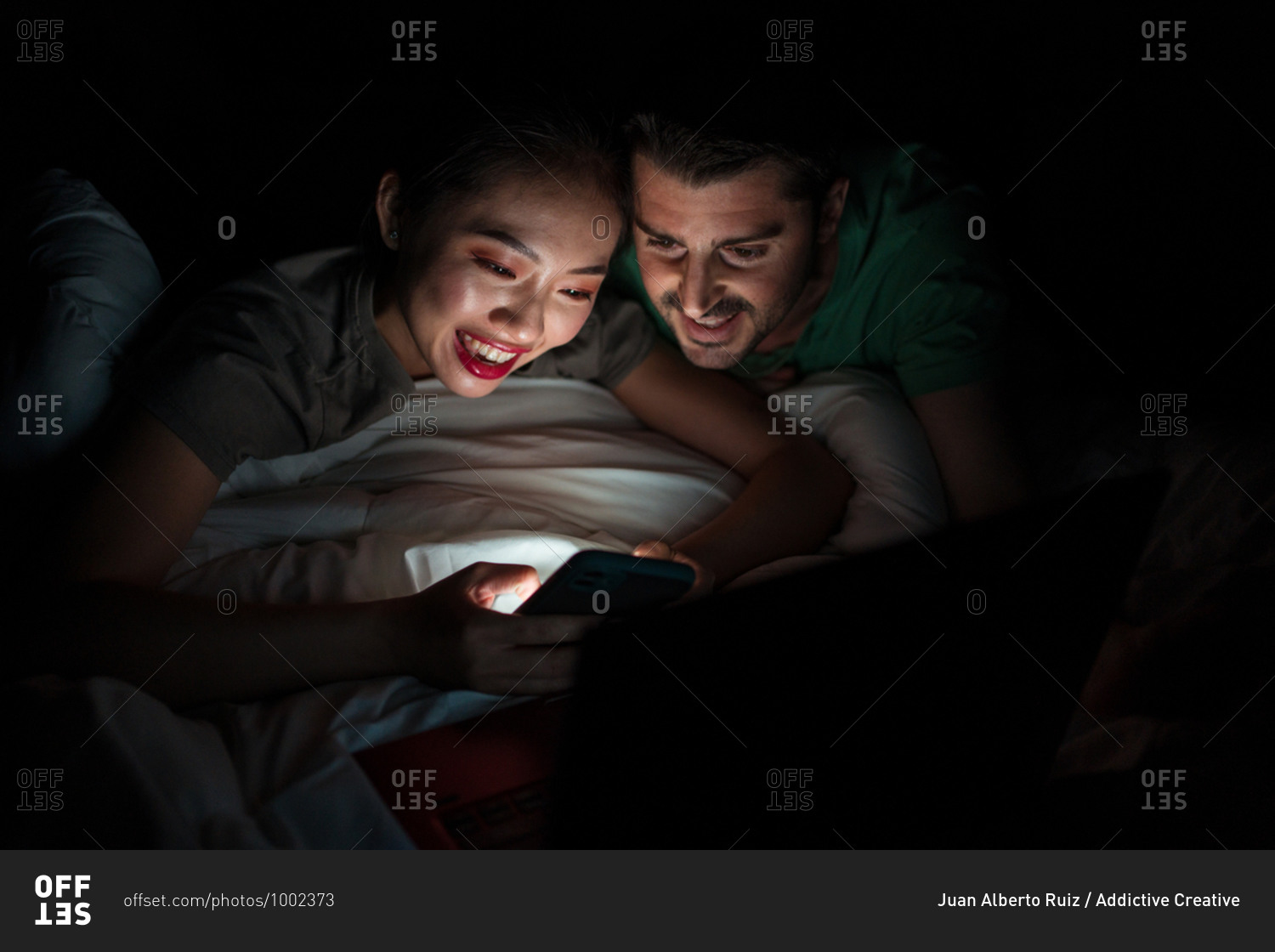 Cheerful young Asian woman using social media on cellphone while lying with happy boyfriend in bed at night in apartment