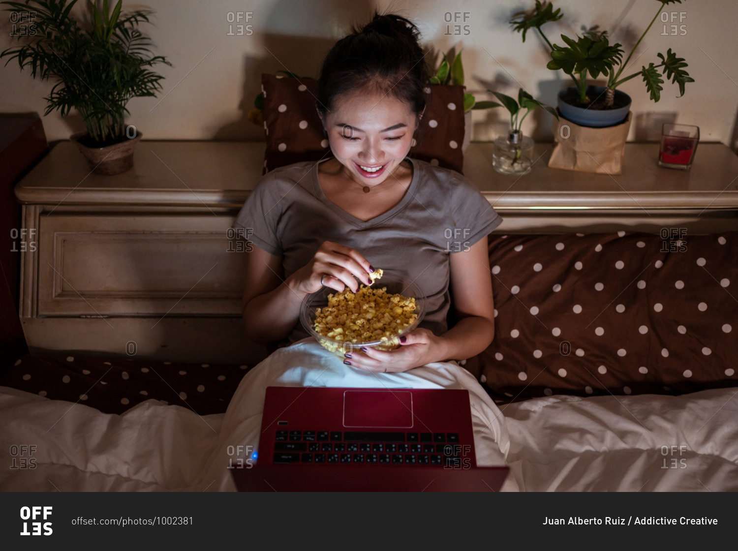 From above of smiling ethnic lady in pajamas sitting on bed with bowl of popcorn while watching film in evening