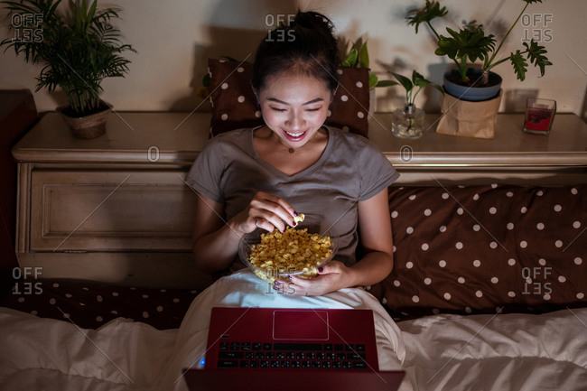 From above of smiling ethnic lady in pajamas sitting on bed with bowl of popcorn while watching film in evening