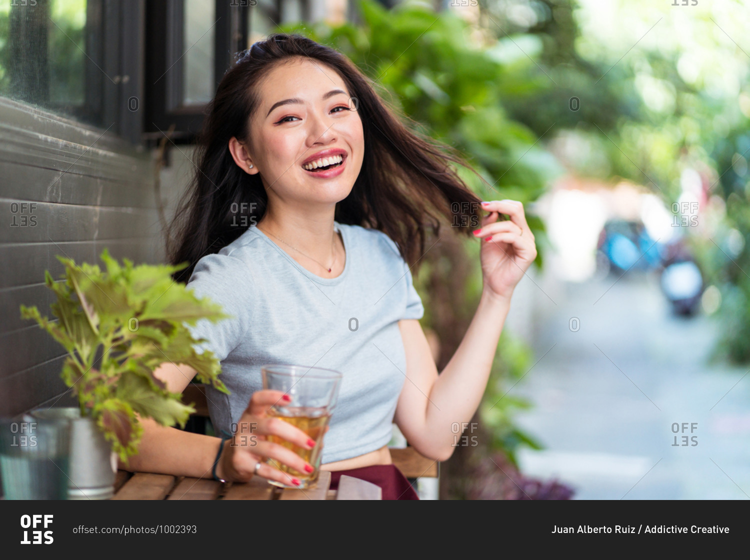 Young Asian female sitting at counter with fresh drink in cafe and enjoying weekend while touching hair and looking at camera