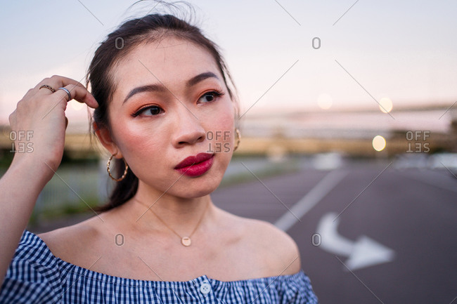 Young thoughtful Asian female in casual wear and golden accessories standing on empty asphalt road with marked arrow under serene sky on blurred background