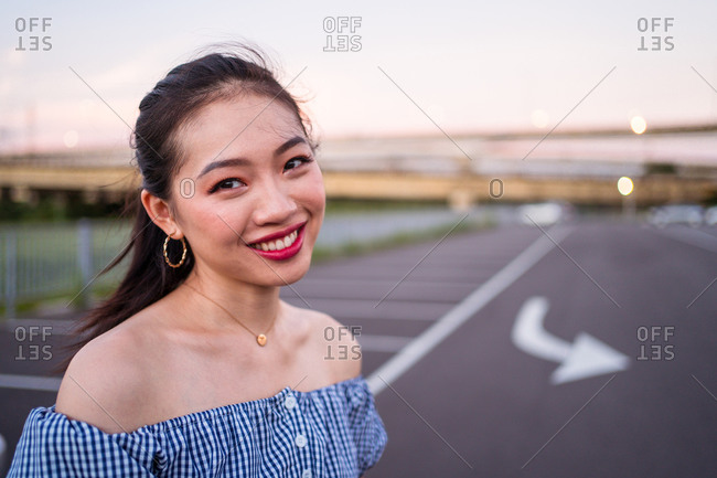 Young cheerful Asian female in casual wear and golden accessories standing on empty asphalt road with marked arrow under serene sky on blurred background