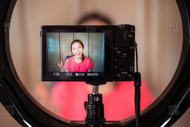 Young Asian female in casual outfit with manicure using camera and ring light while shooting video tutorial for beauty vlog in studio