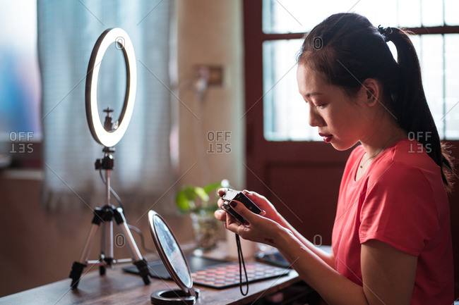 Side view of focused young Asian woman sitting at table with cosmetics and tools for video shooting and checking video on camera while creating content for beauty blog at home