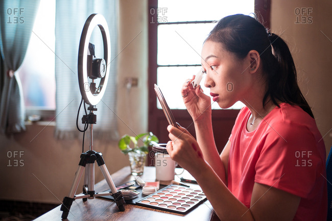 Young Asian woman in casual wear holding mirror and applying foundation on face while recording video tutorial for beauty blog at home workplace with ring light and camera