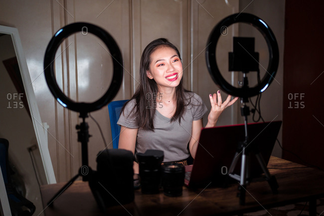 Young Asian female beauty influencer with bright makeup waving hand and talking with subscribers while sitting at table with gadgets and ring light and filming video on camera
