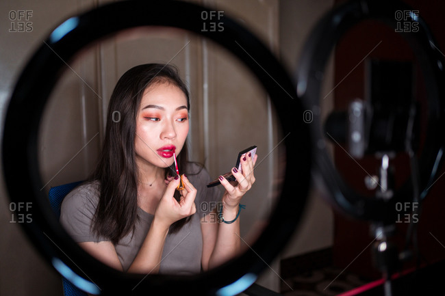 Young Asian woman in casual wear holding mirror and recording video tutorial for beauty blog at home workplace with ring light and camera