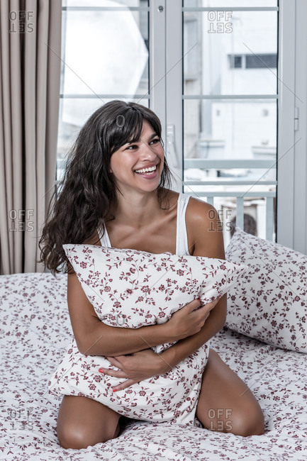 Carefree female sitting on bed hugging pillow while resting and looking away smiling
