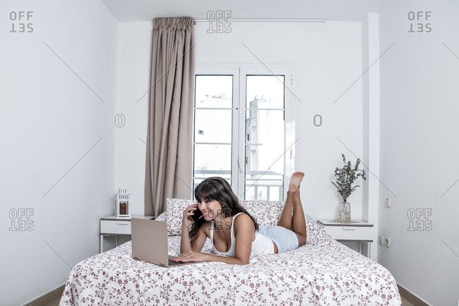 Cheerful female in pajama lying on soft bed using a computer while speaking on the mobile phone enjoying morning and relaxing at home
