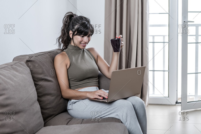 Glad female wearing casual clothing sitting on couch with modern netbook while drinking hot beverage in a mug at light living room