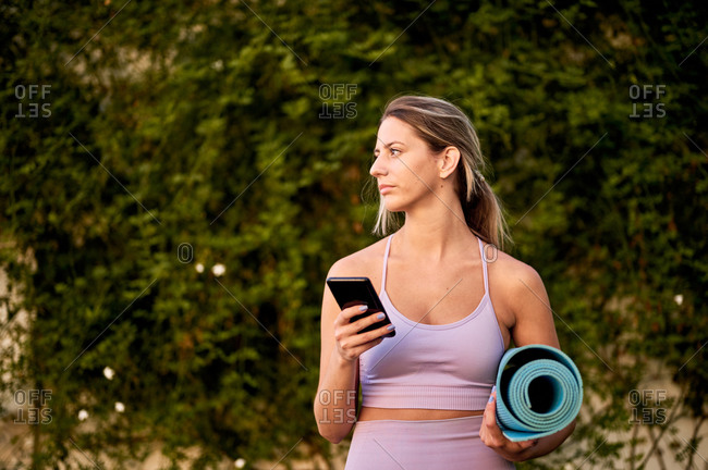 Slim ponder female in sports clothes standing with rolled yoga mat while using cellphone near trees and looking away in daylight
