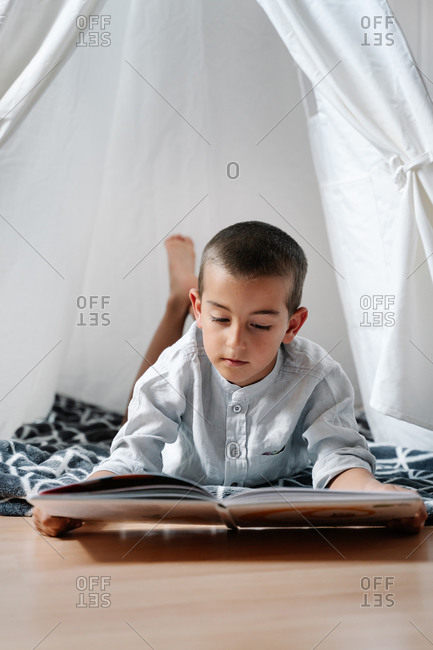 Little boy lying on blanket on wooden floor while reading book in homemade tent decorated with garland of flags in apartment in daylight