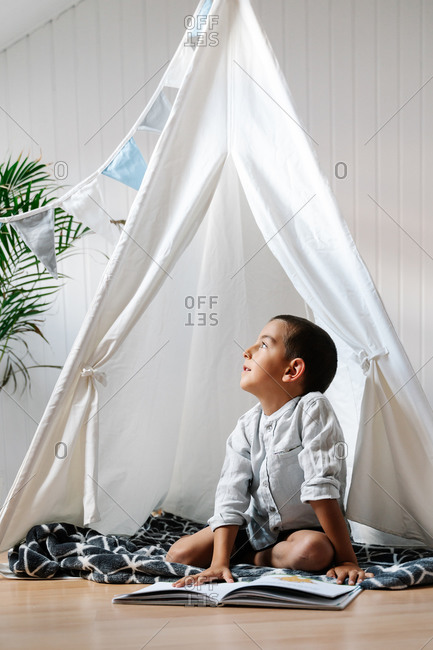 Full body little boy sitting on blanket on wooden floor while reading book in homemade tent decorated with garland of flags in apartment in daylight