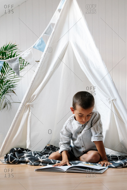 Full body little boy sitting on blanket on wooden floor while reading book in homemade tent decorated with garland of flags in apartment in daylight
