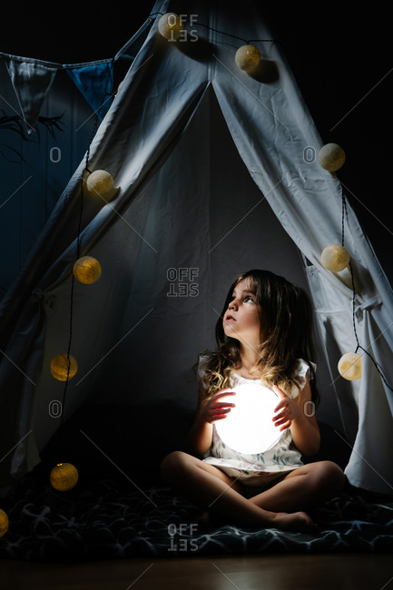 Full body female kid with long hair in casual dress holding illuminating lamp in ball shape while sitting alone on blanket in homemade tent decorated with garland and flags at night