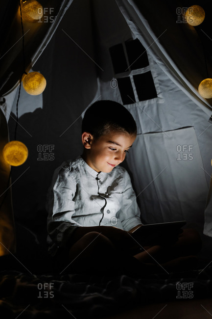 Full length happy boy in casual clothes sitting on blanket watching video on tablet in homemade tent decorated with garland with big lights in moon shapes in darn room at night