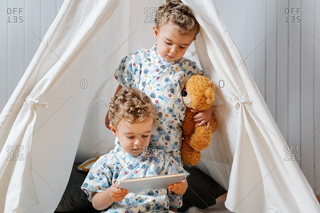 Adorable little twin brothers in similar clothes watching video on tablet while sitting under tent with toys in cozy nursery at home