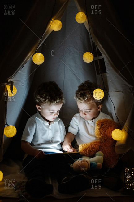 Cute little twin boys with curly hair sitting inside cozy tent with light garland and playing with tablet while spending time together in cozy playroom at home