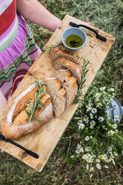 Top view of crop anonymous female holding wooden cutting board with tasty bread cut on loafs with tender texture decorated with fresh rosemary sprigs near bowl with olive oil