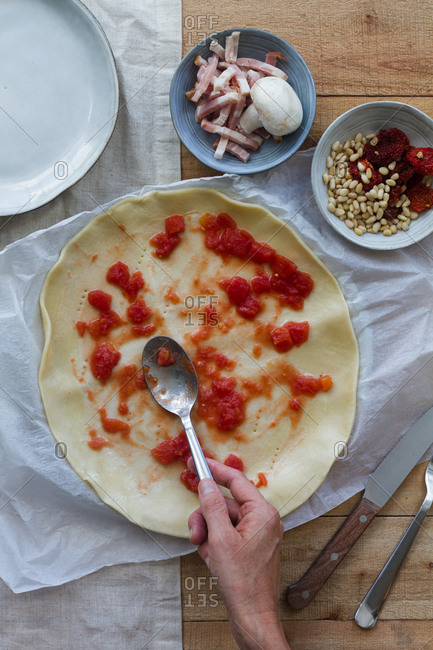 Top view of crop anonymous cook using spoon while putting tomato sauce on raw puff pastry near bowls with bacon slices and pine nuts for quiche during cooking process