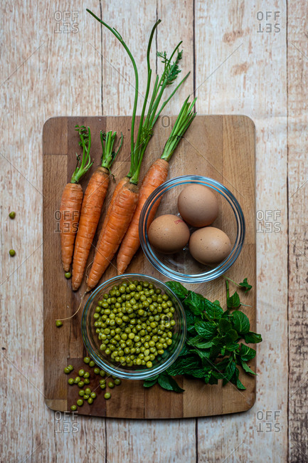 Top view of ripe carrots and basil arranged with canned peas and boiled eggs on wooden chopping board in kitchen