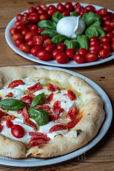 Round dish with yummy Pizza Margherita decorated with basil and dish with small oval tomatoes and basil placed on wooden table in Italian pizzeria