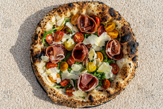 Top view of delicious Italian pizza with small tomatoes and charred cornicione served with prosciutto and cheese decorated with basil leaves placed on concrete background
