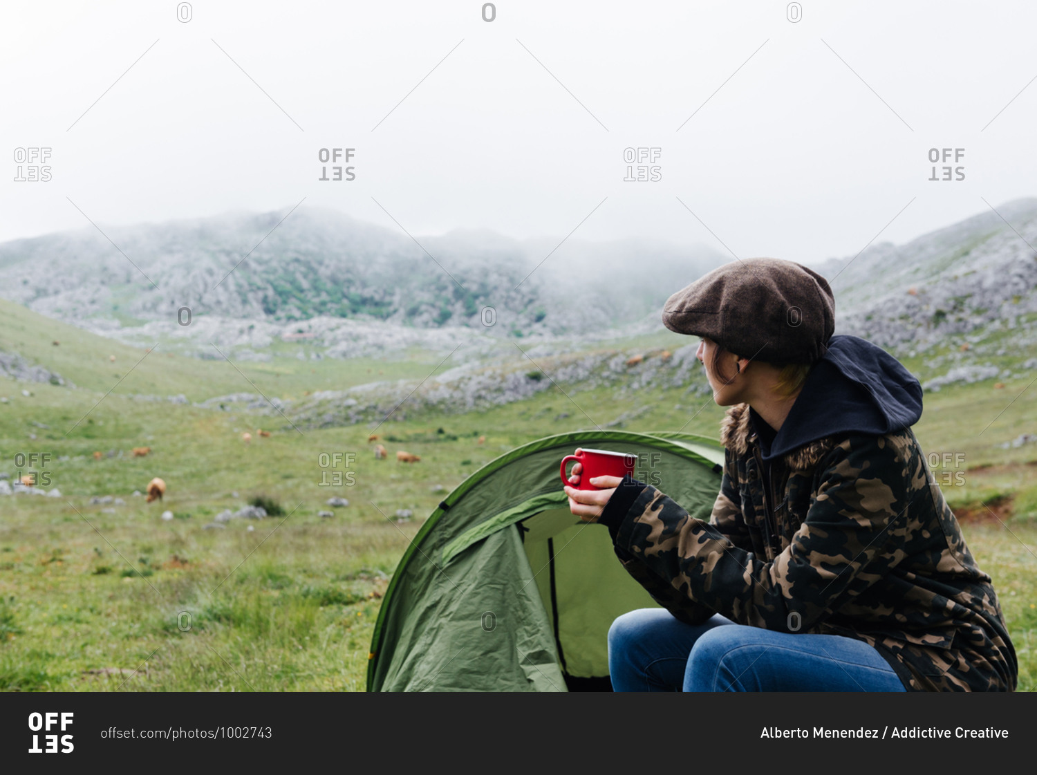 Full length content female traveler in jeans and warm khaki jacket sitting on folding chair near camping tent with cup of hot drink during camping in highlands looking away