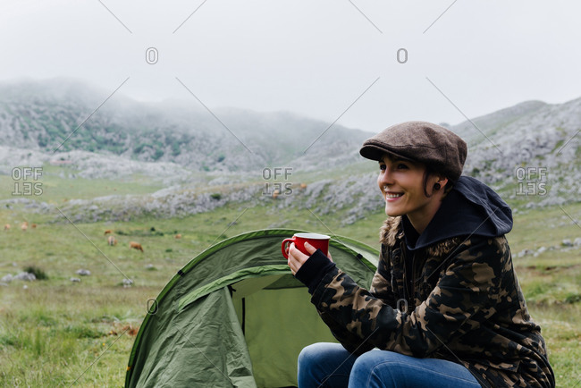 Full length content female traveler in jeans and warm khaki jacket sitting on folding chair near camping tent with cup of hot drink during camping in highlands