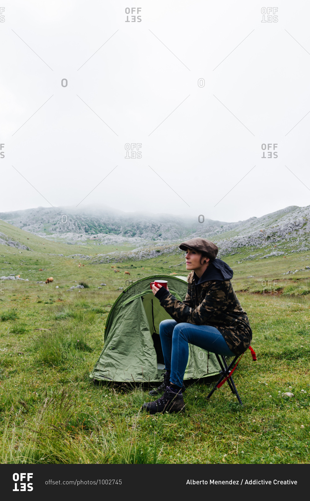 Full length content female traveler in jeans and warm khaki jacket sitting on folding chair near camping tent with cup of hot drink during camping in highlands looking away