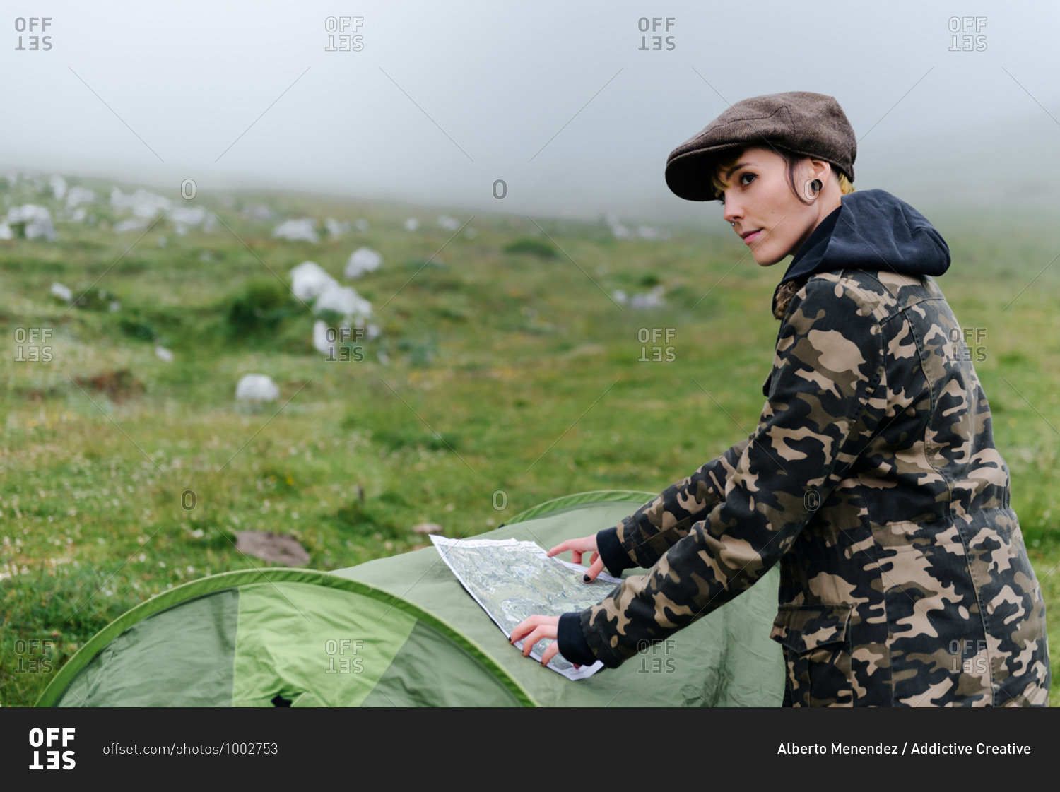 Side view positive female tourist in warm clothes and cap putting map on camping tent and navigating in location while looking away in foggy morning